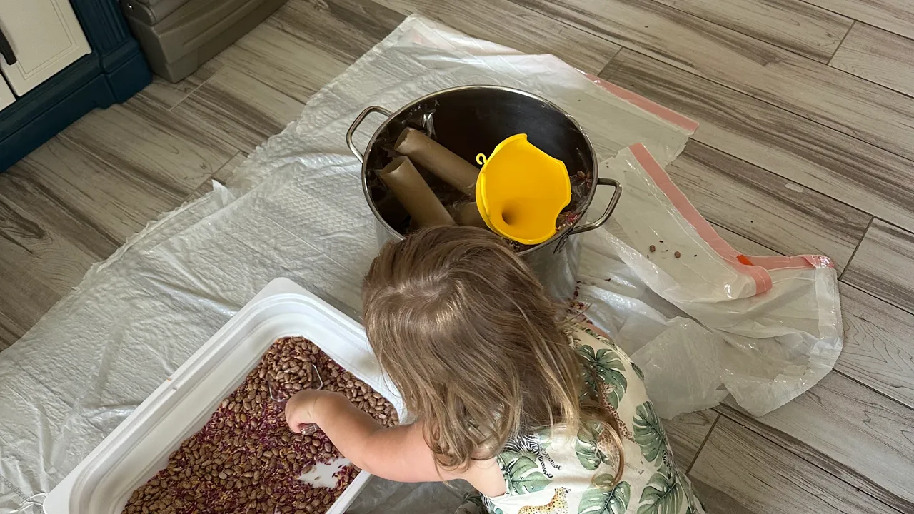 Toddler scooping dried beans through a funnel into a paper towel roll standing upright inside a large pot