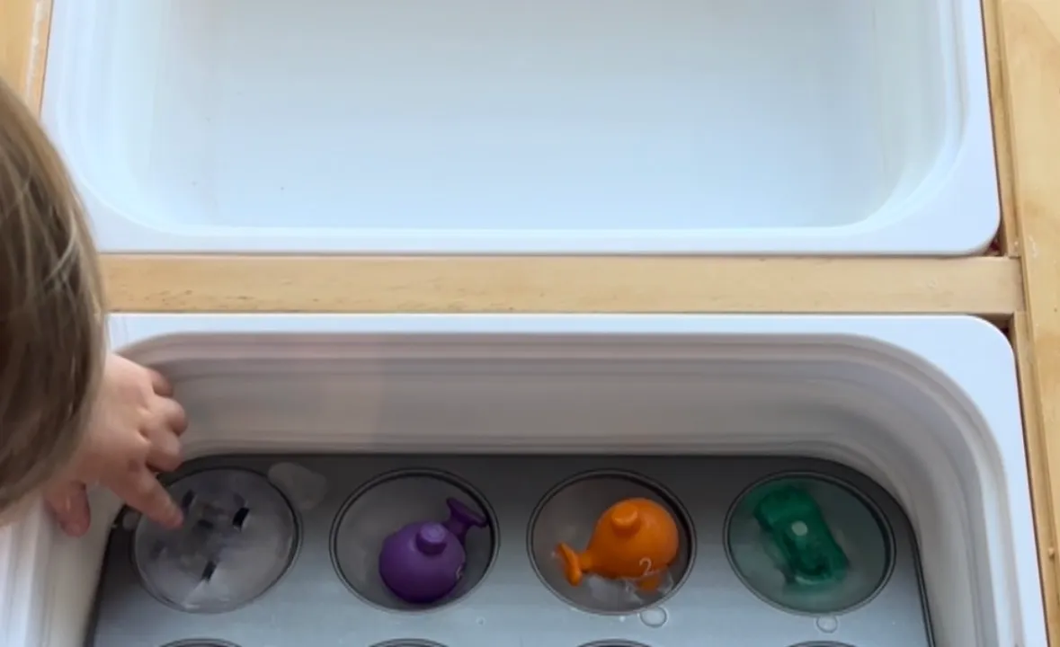 Toddler chipping small plastic toys out of a frozen block of ice sitting in a sensory bin