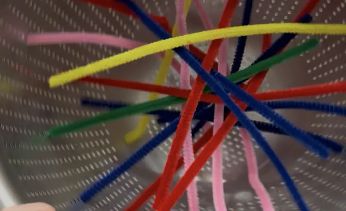 Toddler threading a colored pipe cleaner through the holes of an upside-down metal colander