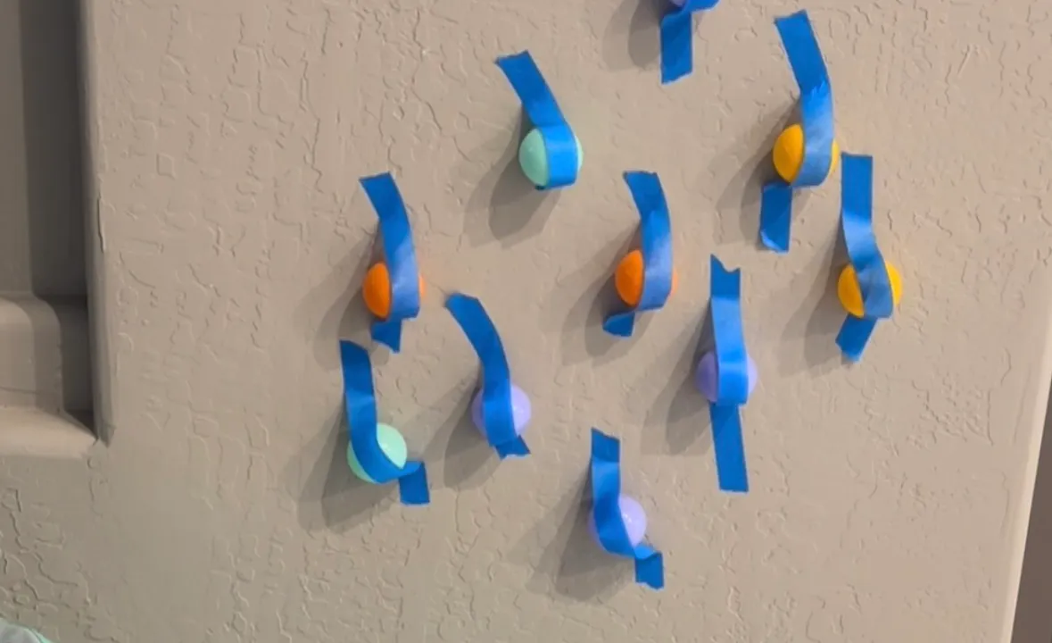 Toddler pulling plastic ball pit balls off strips of painters tape stuck across a wall