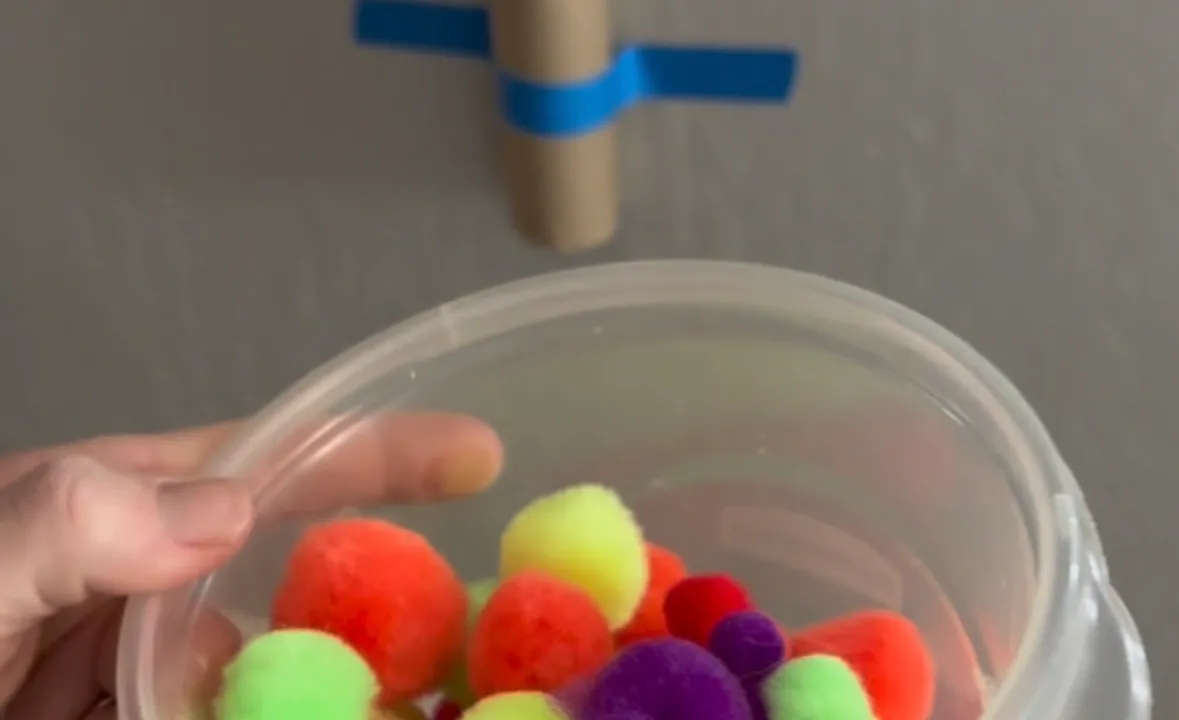 Toddler dropping a pom pom into the open top of a cardboard tube taped to a wall