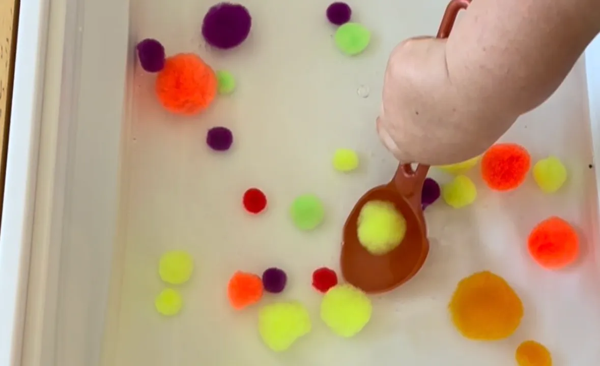Toddler scooping colored pom poms out of a bin of water with a small cup