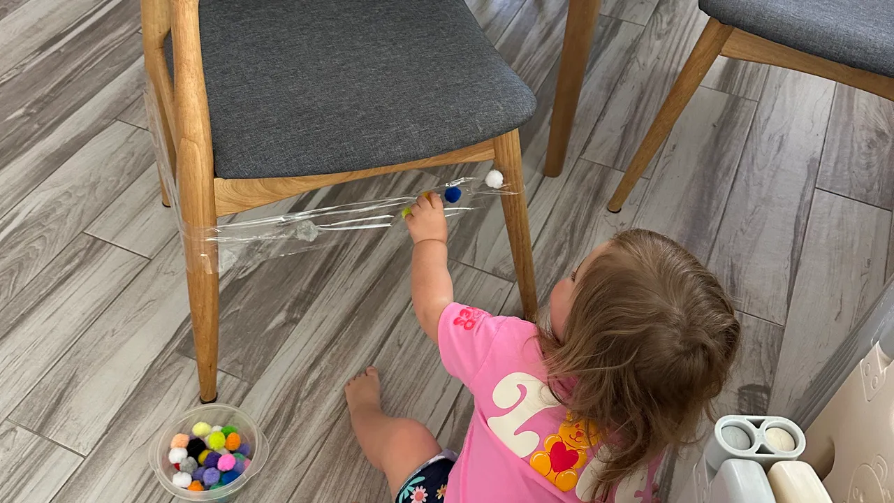 Toddler pressing red and yellow pom poms onto clear packing tape wrapped around a wooden chair leg