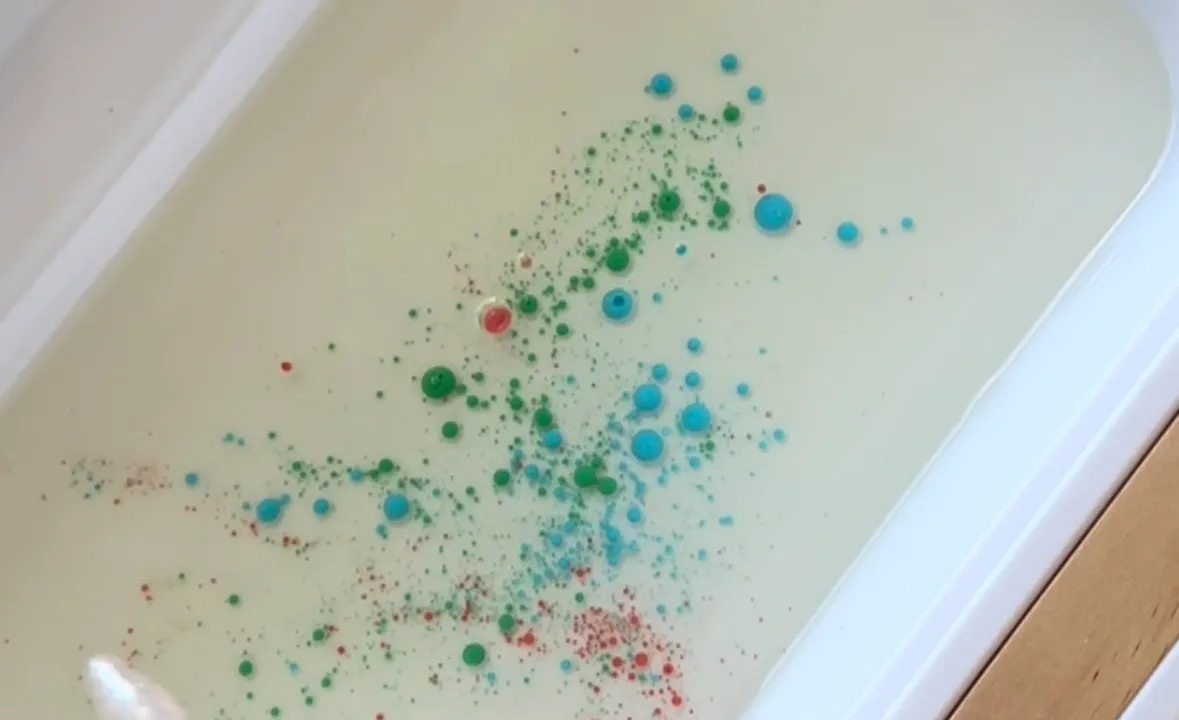 Blue, green and red milk droplets suspended in a white bin of cooking oil while a toddler holds a pink pipette